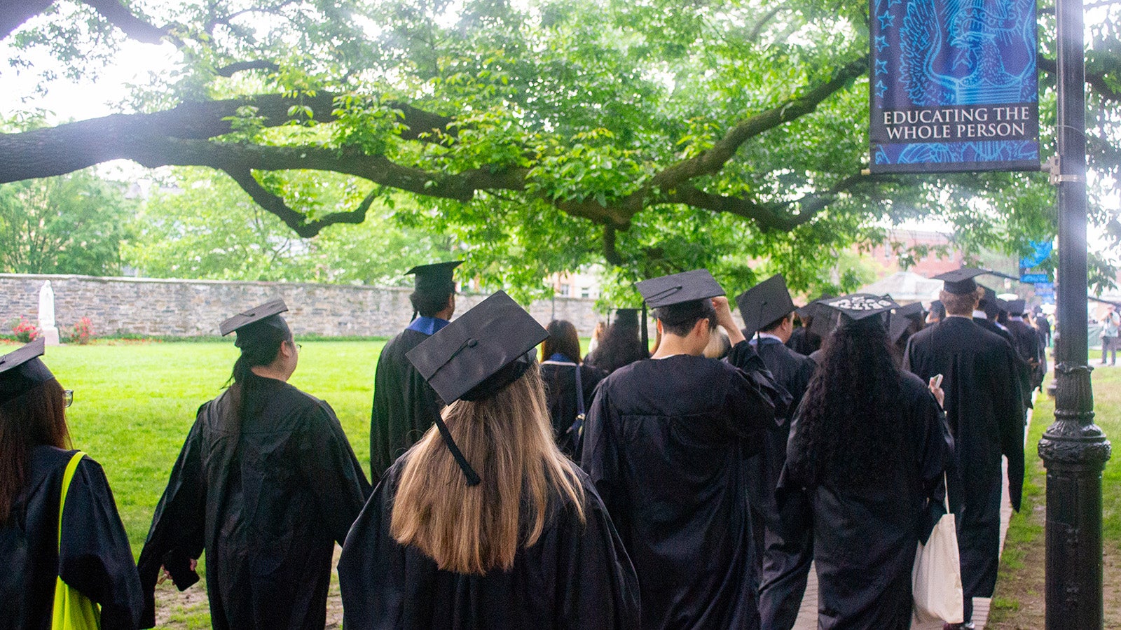 Graduates wearing their regalia process toward the Commencement tent under a Georgetown banner with the words 